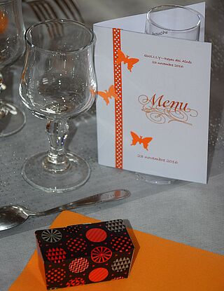 Serviette de table orange avec une petite cuillère et un verre à pied en haut. Sur la serviette, un petit ballotin de chocolat est posé et à droite du verre: un menu en papier est posé debout