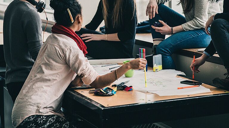 4 jeunes femmes (1 assise sur une chaise, 2 assises sur un mur et 1 assise sur la table) et 1 jeune homme debout en train de travailler autour d'une table où sont posés: des grandes feuilles blanches, des crayons feutres de toutes les couleurs et un téléphone portable