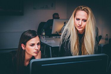 Au 1er plan: 2 femmes regardent un écran d'ordinateur: 1 brune assise et une blonde debout. En arrière-plan, un homme est assis devant un bureau où est posé un ordinateur