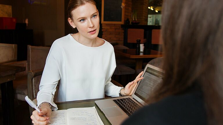 2 femmes face à face: 1 de dos en 1er plan avec les cheveux bruns, 1 avec les cheveux blonds attachés, en train de parler, un stylo à la main pour écrire sur un cahier et un ordinateur portable ouvert entre les 2 femmes