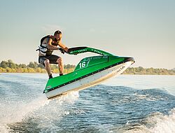 homme en short et gilet de secours saute en jetski vert et blanc au dessus d'un plan d'eau