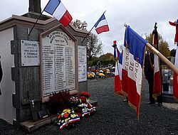 drapeaux français face au monument aux morts du cimetière du Quilly et gerbes de fleurs au pied